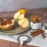 Apfel-Zimt Konfitüre von DELIGREECE mit frischen Apfelscheiben, Zimtstangen und bestrichenem Brötchen auf Teller.