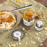 Joghurt mit Müsli und Bergamotte-Orange-Marmelade von DELIGREECE, daneben ein geöffnetes Marmeladenglas und ein Mini-Glas auf einem mit Nüssen bestreuten Tuch.