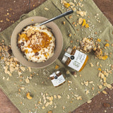 Schale mit Joghurt und Müsli, verfeinert mit Bergamotte-Orange-Marmelade von DELIGREECE; daneben zwei Marmeladengläser auf einem Tuch mit verstreuten Nüssen und Getreideflocken.