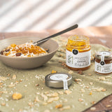 Schale mit Müsli und Joghurt, verfeinert mit Bergamotte-Orange-Marmelade von DELIGREECE; daneben zwei Marmeladengläser und ein Deckel auf einem mit Nüssen und Haferflocken bestreuten Tuc