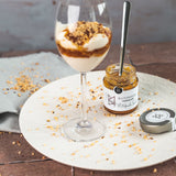 Joghurt-Dessert im Weinglas mit Schichten aus Blutorangen-Marmelade von DELIGREECE, garniert mit gehackten Nüssen; daneben das geöffnete Marmeladenglas mit Löffel und Deckel auf einem hellen Teller