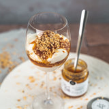 Dessert im Weinglas mit griechischem Joghurt, karamelliger Pfirsich-Konfitüre von DELIGREECE und gehackten Nüssen, daneben das geöffnete Marmeladenglas.