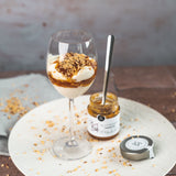 Weinglas mit Joghurt-Dessert, verfeinert mit Blutorangen-Marmelade von DELIGREECE und gehackten Nüssen, daneben das geöffnete Marmeladenglas und der Deckel.
