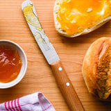 Marmeladenmesser mit Holzgriff liegt auf einem Holzbrett neben Brötchen und Schale mit Marmelade.
