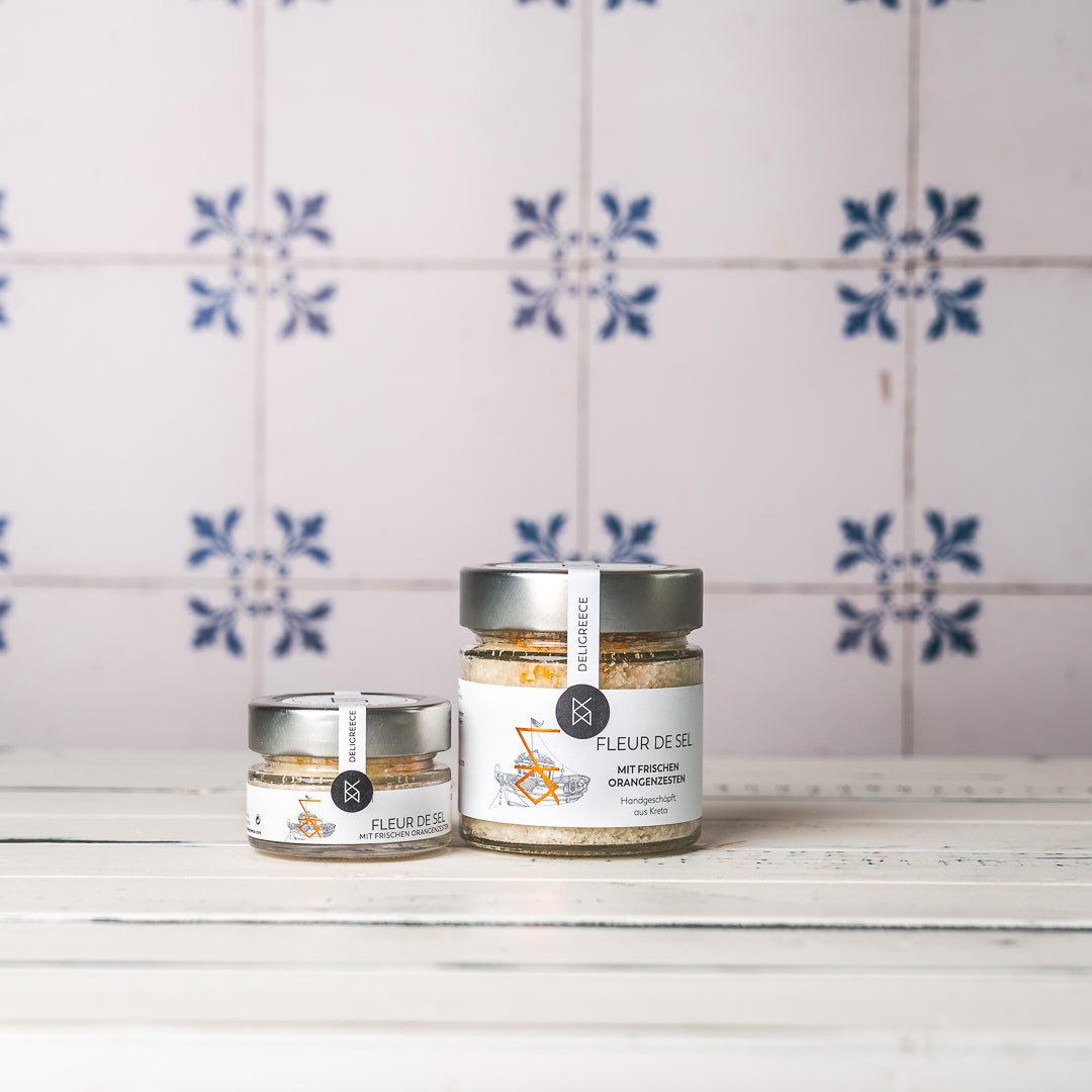 DELIGREECE Fleur de Sel mit frischen Orangenzesten – zwei Gläser in unterschiedlichen Größen auf weißem Holztisch, vor hellem Hintergrund mit blauen Fliesenmotiven.