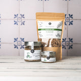 DELIGREECE Fleur de Sel mit kretischen Wildkräutern – zwei Gläser und eine große Nachfüllpackung auf weißem Holztisch vor hellen Fliesen mit blauem Muster.