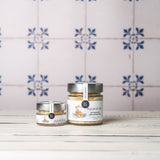 DELIGREECE Fleur de Sel mit frischen Orangenzesten – zwei Gläser in unterschiedlichen Größen auf weißem Holztisch, vor hellem Hintergrund mit blauen Fliesenmotiven.