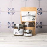 DELIGREECE Fleur de Sel – geräuchertes Meersalz aus Kreta in zwei Gläsern und einer großen Nachfüllpackung, arrangiert auf weißem Holztisch vor hellen Fliesen mit blauem Muster.