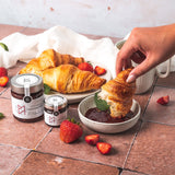 Erdbeer-Konfitüre von DELIGREECE mit Croissants, frischen Erdbeeren und einer Hand, die ein Croissant in die Konfitüre tauch