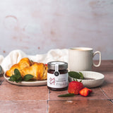 Erdbeer-Konfitüre von DELIGREECE mit Croissants, Erdbeeren, Minze und einer Tasse Kaffee auf rustikalem Untergrund.