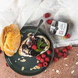 Frühstücksszene mit Brot, Frischkäse und Himbeerkonfitüre von DELIGREECE auf Teller, dekoriert mit frischen Himbeeren und Mandelblättchen; daneben das Himbeer-Konfitüre-Glas.