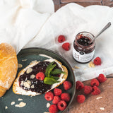 Scheibe Brot mit Frischkäse und Himbeerkonfitüre von DELIGREECE auf einem Teller, garniert mit frischen Himbeeren und Minze; daneben geöffnetes Himbeer-Konfitüre-Glas und verstreute Mandelblättchen.