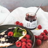 Geöffnetes Himbeer-Konfitüre-Glas von DELIGREECE mit Löffel, daneben frische Himbeeren und eine bestrichene Brotscheibe auf Teller.