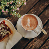 Heißer Kakao als Servierbeispiel – cremiger Kakao mit Latte Art in einer Tasse, serviert auf Holz mit Kuchenstück daneben.