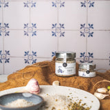 DELIGREECE Fleur de Sel Knoblauch & Kräuter – zwei Gläser auf braunem Naturtuch arrangiert, umgeben von Knoblauchzehen und Gewürzen, vor traditioneller Fliesenwand.
