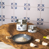 Fleur de Sel Knoblauch & Kräuter von DELIGREECE, zwei Gläser auf braunem Tuch arrangiert, dazu Knoblauchzehen und eine Schale mit Salzmischung vor traditioneller Fliesenwand.