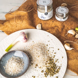 Fleur de Sel mit Wildkräutern aus Kreta, angerichtet auf einem weißen Teller mit frischem Knoblauch und Kräutern, daneben zwei DELIGREECE Salzgläser auf braunem Tuch.