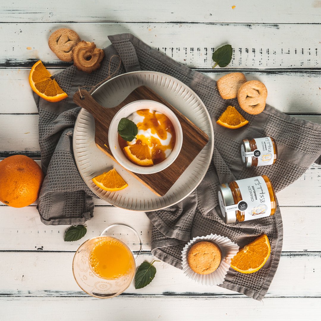 Orangen-Konfitüre Extra von DELIGREECE, serviert mit Joghurt, Orangenstücken, Gebäck und frischem Saft auf hellen Holzbrettern.