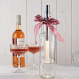 dekorative Glasflasche mit Bottlelight-LED im Inneren und rot-weiß karierter Schleife am Flaschenhals; im Hintergrund eine Roséweinflasche und zwei gefüllte Weingläser auf hellem Tisch vor weißer Holzpaneel-Wand.