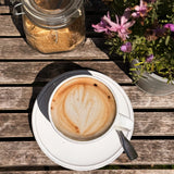 Dinkel Cappuccino in Tasse auf Holztisch im Freien, neben Blumen und Glas mit Pulver