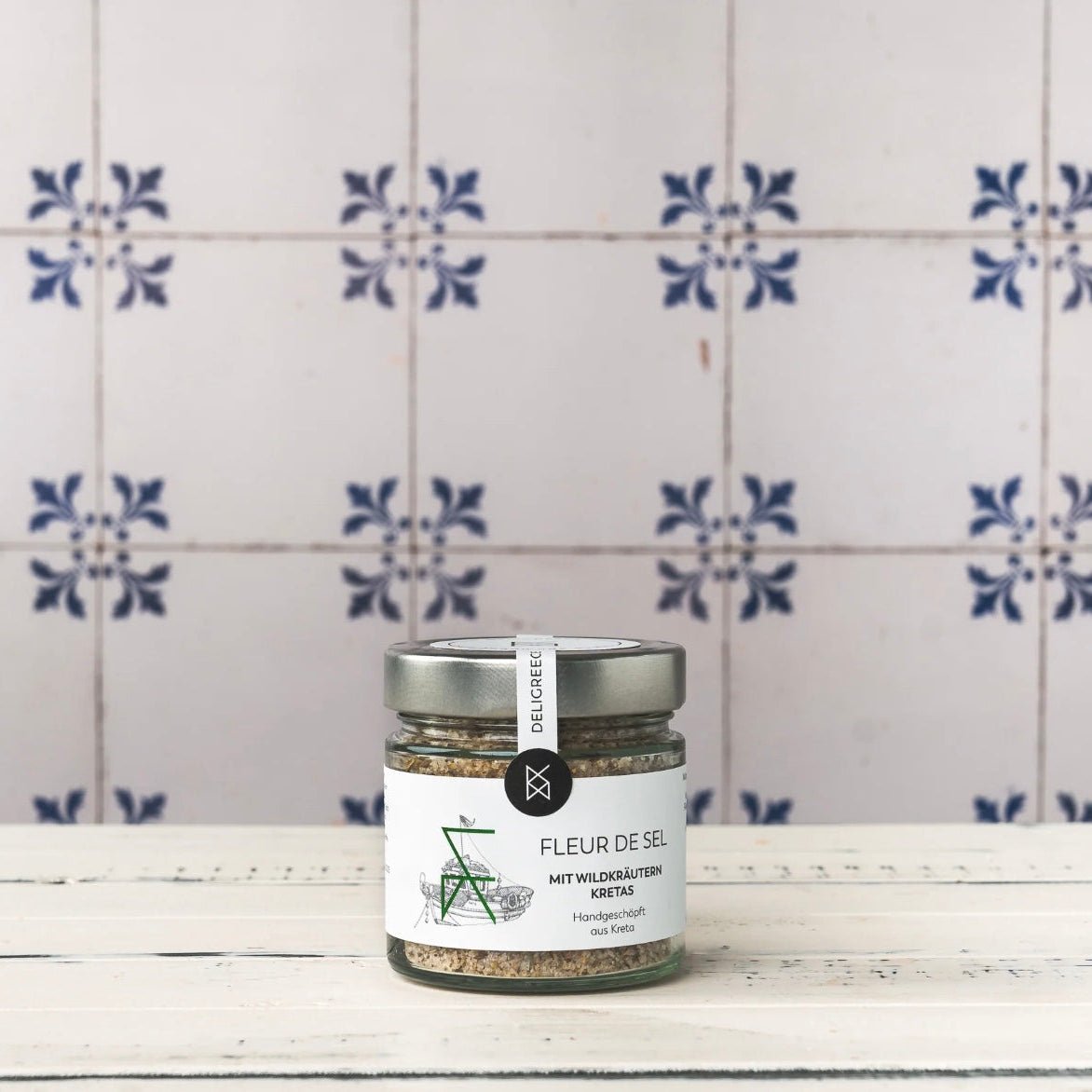 Glas Fleur de Sel mit Wildkräutern (Kreta) auf weißem Holztisch vor blau-weiß gefliester Wand