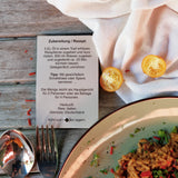 Rezeptkarte mit der Überschrift ‚Zubereitung / Rezept‘ neben einem Teller Reisgericht; daneben Besteck, halbierte gelbe Tomaten und ein helles Tuch auf einem Holztisch.